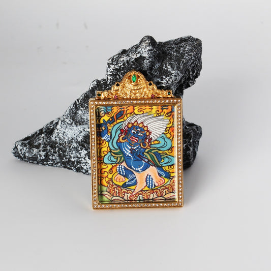 Angled view of Acalanatha (Fudo Myo-o) mini thangka pendant showing the hand-painted blue wrathful deity figure with flame background, gold-tone rectangular frame with crystal border, and ornate crown top with a green gemstone, photographed on a rock prop.