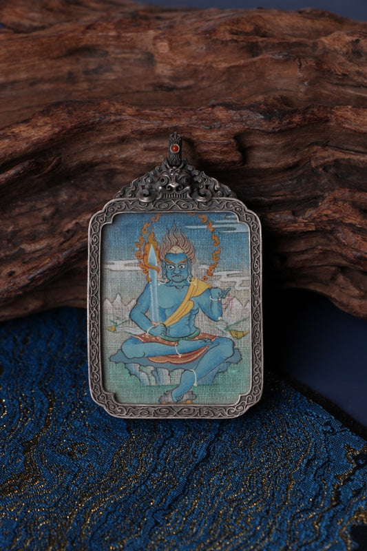 Close-up of an Acalanatha (Fudo Myo-o) mini thangka pendant, hand-painted with mineral pigments and framed in a detailed vintage silver gawu box, photographed on a wooden surface with blue fabric.