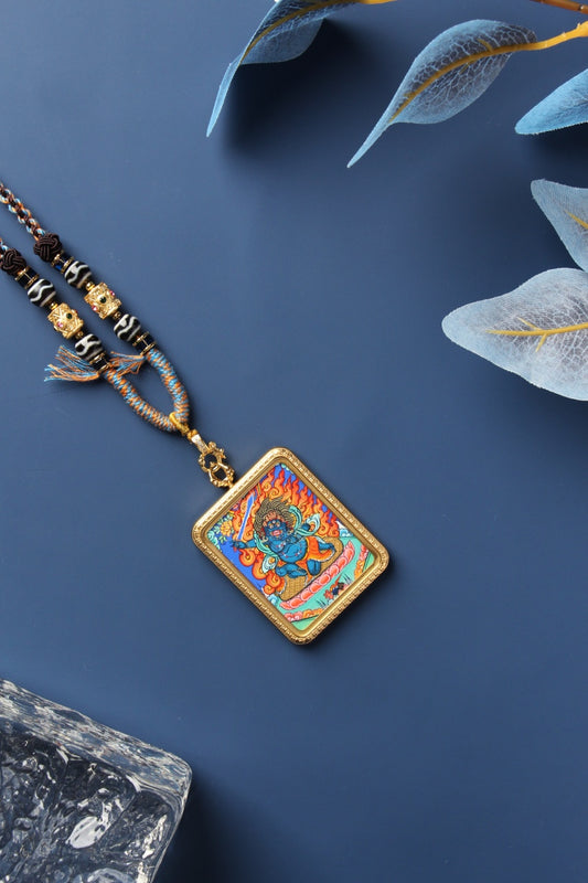Diagonal flat-lay photo of an Acalanatha (Fudo Myo-o) mini thangka pendant with a gold-tone titanium steel case, braided cord, and bead details on a dark blue background.