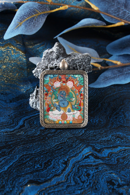 Front view of an Acalanatha (Fudo Myo-o) Menri-style mini thangka pendant, hand-painted in mineral pigments and set in a silver gawu case on blue fabric with stone.