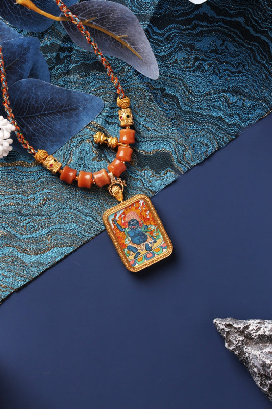 Acalanatha mini thangka pendant necklace showing a hand-painted wrathful blue deity with flame halo inside a rectangular metal frame, paired with braided cord and orange beads