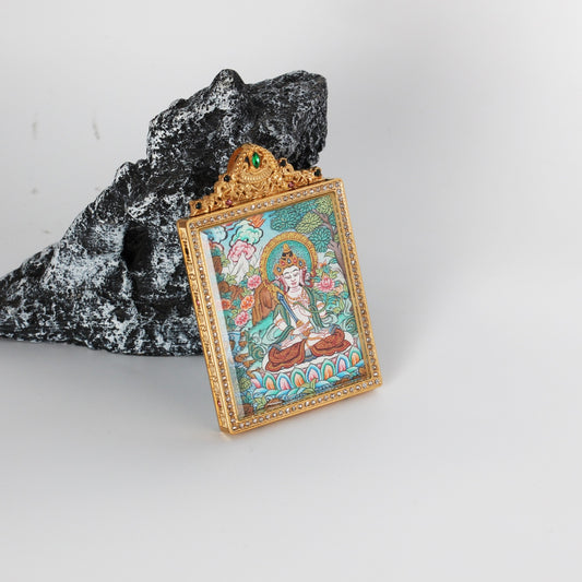 Angled view of Akashagarbha Bodhisattva mini thangka pendant in a gold-tone jeweled frame, set against a rock backdrop.