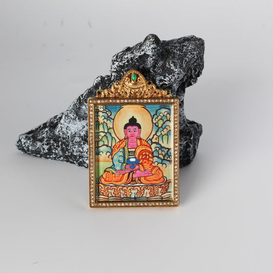 Front view of Amitabha Buddha mini thangka pendant showing the hand-painted seated Amitabha Buddha with halo, gold-tone rectangular frame with crystal border, and ornate crown top with a green gemstone, photographed on a rock prop.