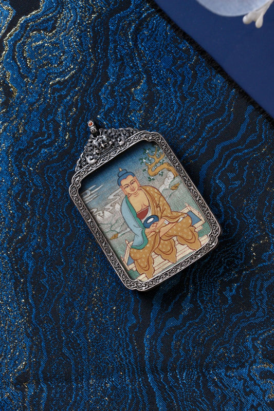 Hand-painted Amitabha Buddha mini thangka set in an ornate vintage silver gawu pendant, photographed on blue brocade textile.