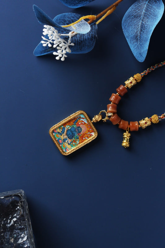 Angled view of Acalanatha thangka pendant highlighting the rectangular framed painting, braided cord necklace, orange bead strand, and small metal prayer wheel charm