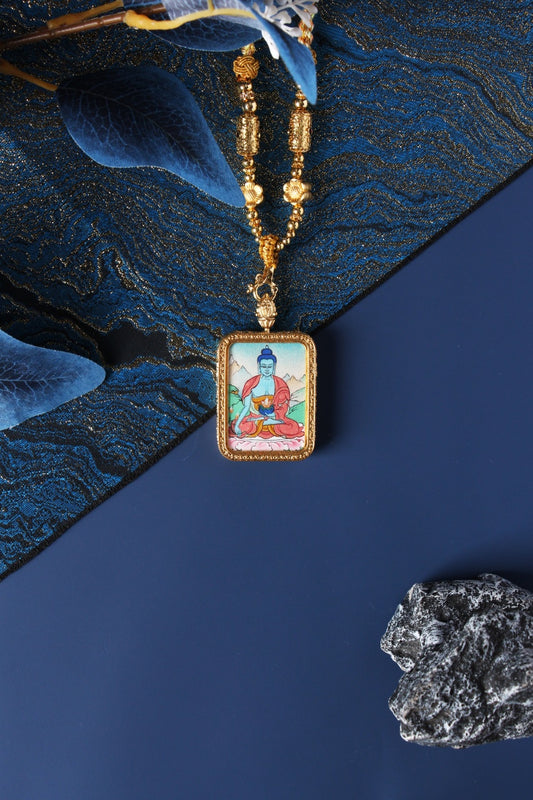 Rectangular Bhaisajyaguru Buddha thangka pendant necklace in a gold-tone frame on a dark blue background.