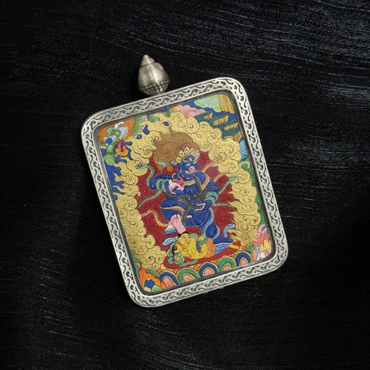 Angled view of a Black Jambhala thangka pendant set in an engraved silver gawu frame, showing the blue wrathful wealth deity holding ritual objects, painted with layered mineral pigments and intricate gold flame background.