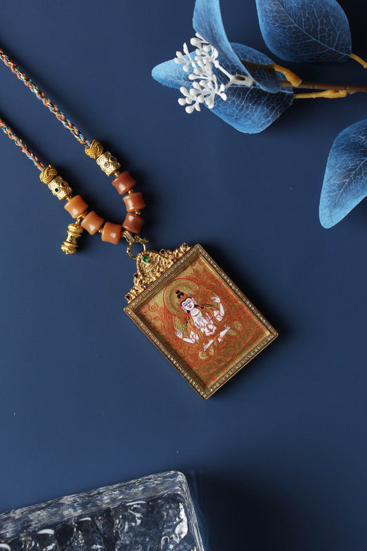 Angled view of a Four-Armed Avalokiteshvara gold thangka pendant showing detailed mineral pigment painting, ornate metal frame, prayer bead elements, and multicolor braided cord.