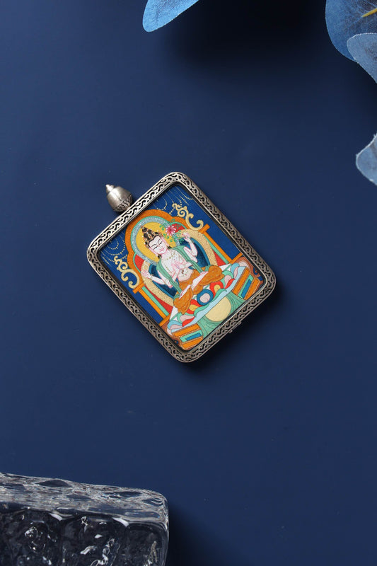 Flat lay of a hand-painted Four-Armed Avalokiteshvara (Chenrezig) mini thangka pendant in a Dunhuang Nine-Color Deer sterling silver case on a dark blue background