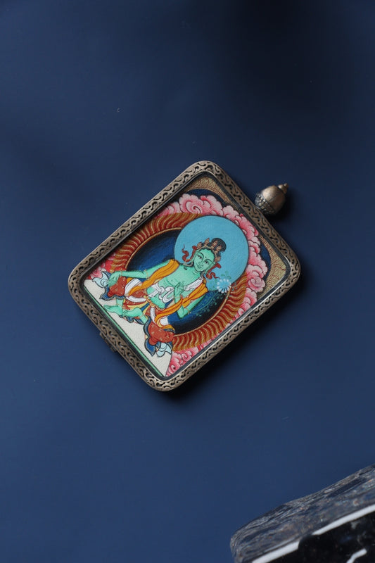 Angled product view of the Green Tara mini thangka pendant from the Tibetan Red Cloud Zangka Series, fully hand-painted in Tibet with natural mineral pigments and presented in a Dunhuang Nine-Colored Deer sterling silver case for portable devotion and collecting.