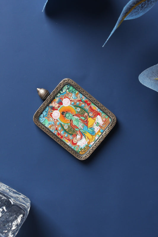 Angled display of a Green Tara mini thangka pendant from Tibet, Menri style painting in mineral pigments, framed in a Dunhuang Nine-Color Deer silver case on a dark blue background.