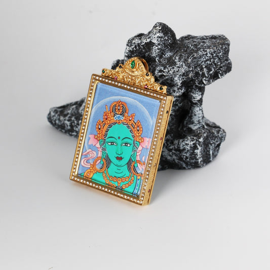 Angled view of Green Tara Buddha head mini thangka pendant showing the raised titanium steel housing, crystal-style bezel border, and decorative crown-like top with colored accents