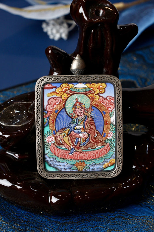 Close-up of a hand-painted Guru Rinpoche seated mini thangka pendant set in a silver gawu case on a dark display stand.
