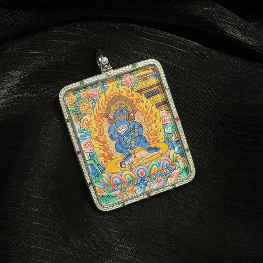 Close-up of a hand-painted Black Jambhala mini thangka pendant showing the blue wrathful wealth deity seated on a lotus base surrounded by flames, floral motifs, and Tibetan architectural elements.