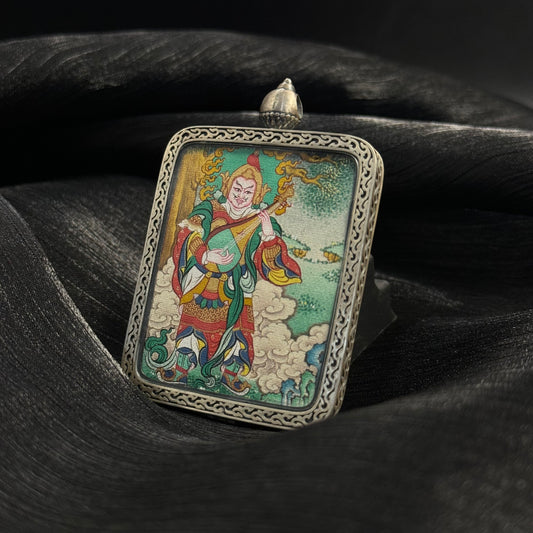 Angled perspective of Dhṛtarāṣṭra Eastern Guardian King mini thangka pendant showing detailed hand-painted figure in armor holding a pipa, surrounded by clouds and landscape elements inside a carved sterling silver pendant frame.