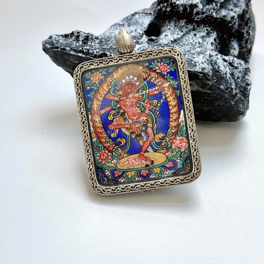 Angled view of Kurukulla (Red Tara manifestation) mini thangka pendant, fully hand-painted with mineral pigments, vivid blue background and floral details, set in a Dunhuang Nine-Color Deer silver case with patterned border