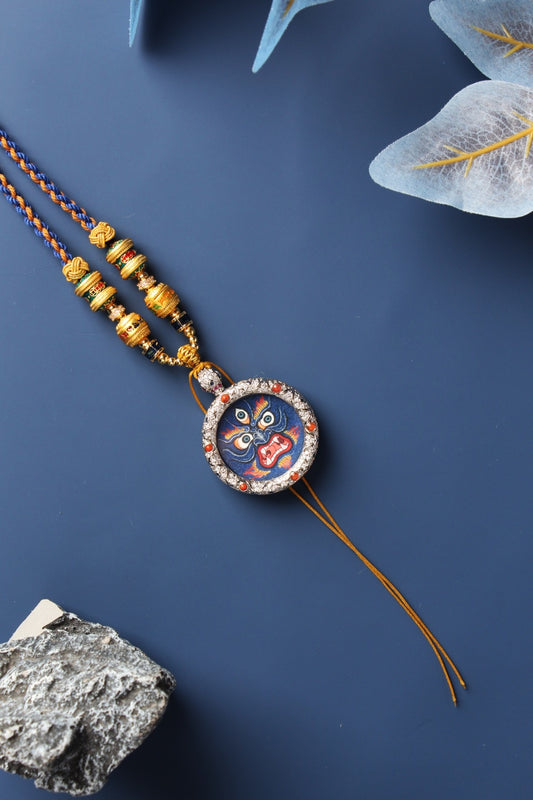 Diagonal flat-lay photo of a Mahakala mini thangka pendant highlighting the intense facial expression, rough mineral pigment texture, copper setting, and traditional beaded necklace.