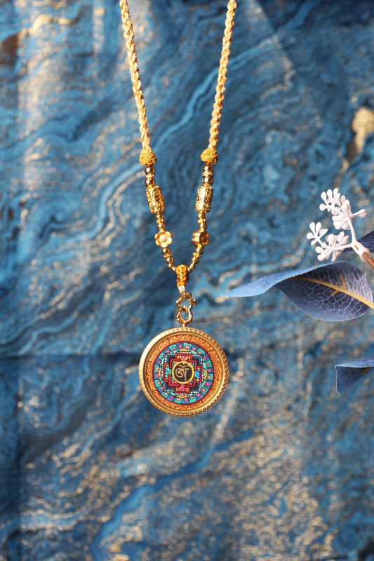Vertical display showing a mandala mini thangka pendant in a round titanium steel case, highlighting the balanced geometric design, braided necklace, and gold-tone bead details against a textured blue background.