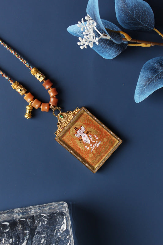 Angled view of Vairocana Buddha mini thangka pendant showing fine gold thangka linework, layered mineral pigment texture, gemstone-accented titanium steel frame, and braided cord connection