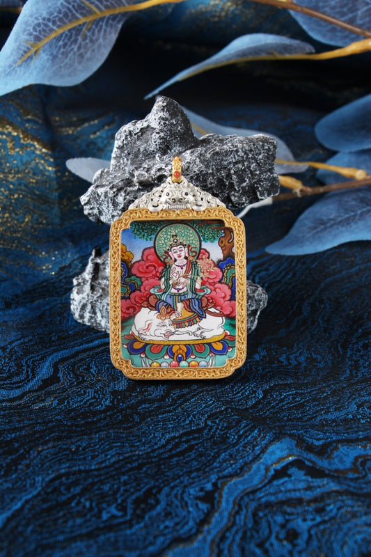 Close-up portrait of Samantabhadra Bodhisattva mini thangka pendant, fully hand-painted with mineral pigments, featuring fine linework and vivid clouds, framed in a gold-tone border with an ornate silver-plated case top, photographed against blue brocade fabric with layered translucent leaves and stone props.