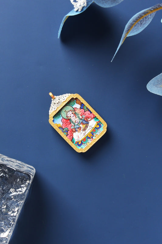 Angled flat lay of Samantabhadra Bodhisattva mini thangka pendant, fully hand-painted with mineral pigments, set in an ornate silver-plated case with gold-tone border and a small red accent at the bail, photographed on a deep navy surface with translucent leaf decor and a textured stone prop.