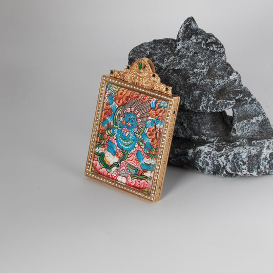 Front view of a hand-painted Six-Armed Mahakala mini thangka pendant, set in a gold-tone jewelry-grade titanium steel case with crystal-set border and green gemstone crown detail, painting size 4 × 5 cm.
