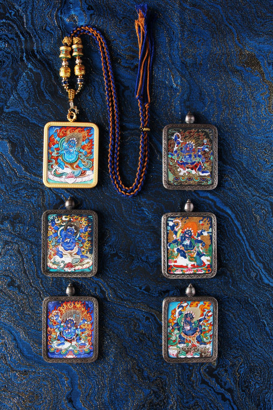 Group display of Six-Armed Mahakala mini thangka pendants with hand-painted mineral pigment artwork, shown in copper cases with braided blue-and-orange cord on a textured blue fabric background.