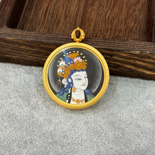 Full view of a hand-painted Sudhana Avalokiteshvara mini thangka pendant on a grey fabric background, showing the round gold-tone frame, hanging loop, and portrait composition.