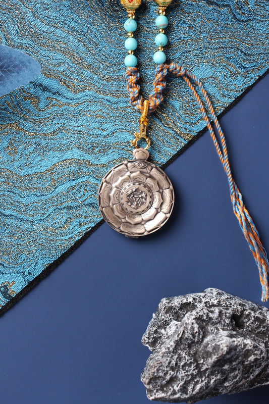 Back view of a round 925 silver pendant case with an engraved floral pattern and raised Three Hares emblem, attached to a braided cord and gold-tone clasp.