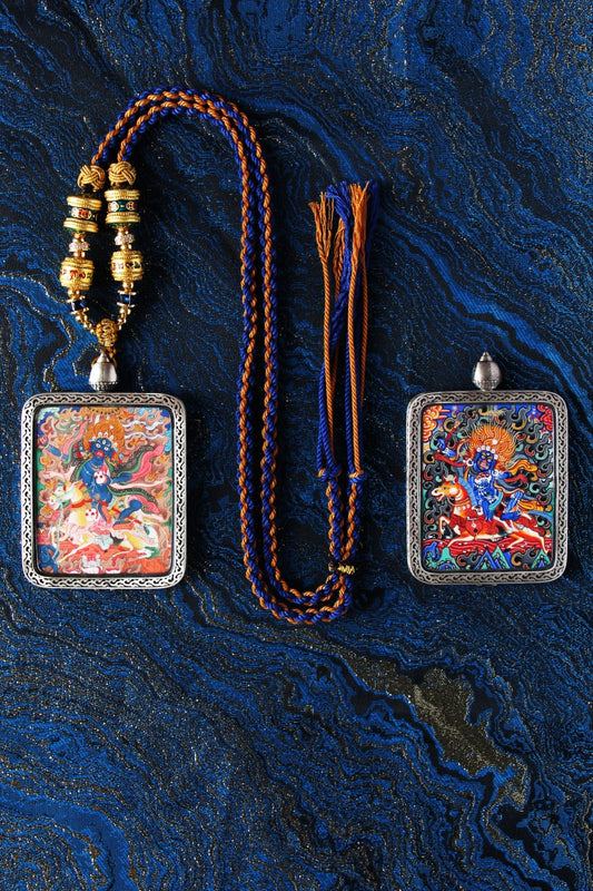 Flat-lay showing two Palden Lhamo mini thangka pendants side by side, each framed in a silver protective case, with a blue-and-gold braided cord necklace and loose tassel cords on a deep blue brocade background