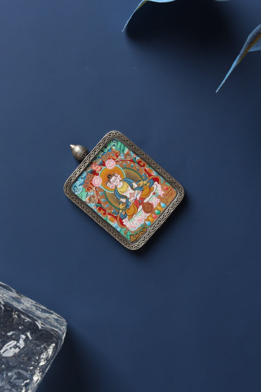 Angled flat lay of a Vairocana Buddha mini thangka pendant in an engraved silver gawu-style case on a dark blue background.
