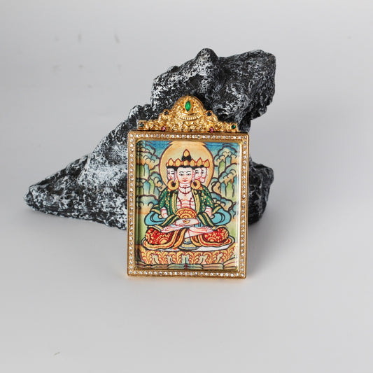 Front view of Vairocana Buddha mini thangka pendant showing the hand-painted multi-faced Vairocana figure with halo, gold-tone rectangular frame with crystal border, and ornate crown top with a green gemstone, photographed on a rock prop.