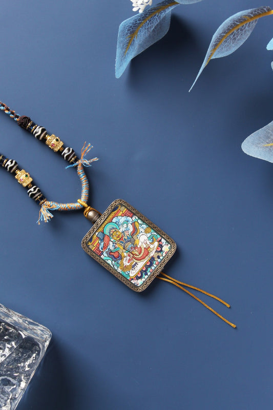 Angled flat-lay of Vaishravana (Bishamonten) mini thangka pendant, hand-painted with mineral pigments in Rebgong style, set in a Dunhuang Nine-Color Deer sterling silver protective case with braided cord and tassel ends