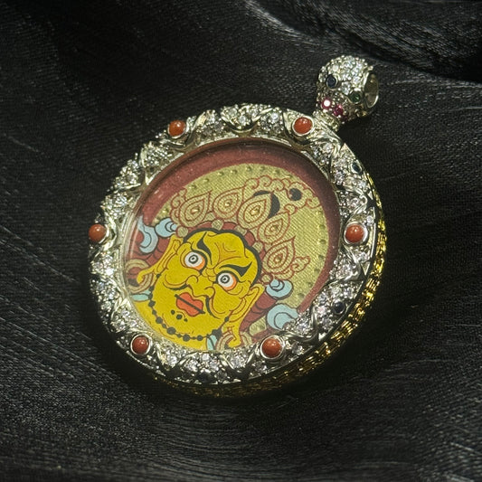 Detailed angled view of Yellow Jambhala mini thangka pendant showing the expressive Rebgong-style deity face, cinnabar red halo background, and decorative gemstone copper frame.