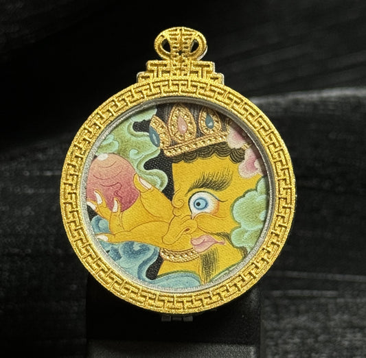 Front view of a circular Yellow Jambhala mini thangka pendant, hand-painted with mineral pigments, showing the side-profile of the Tibetan wealth deity holding a wish-fulfilling jewel inside a gold-tone protective frame.