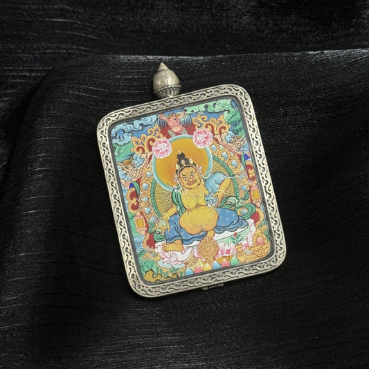 Angled close-up of Yellow Jambhala mini thangka pendant showing layered mineral pigments, detailed facial expression, golden halo, lotus base, and carved silver gawu frame.