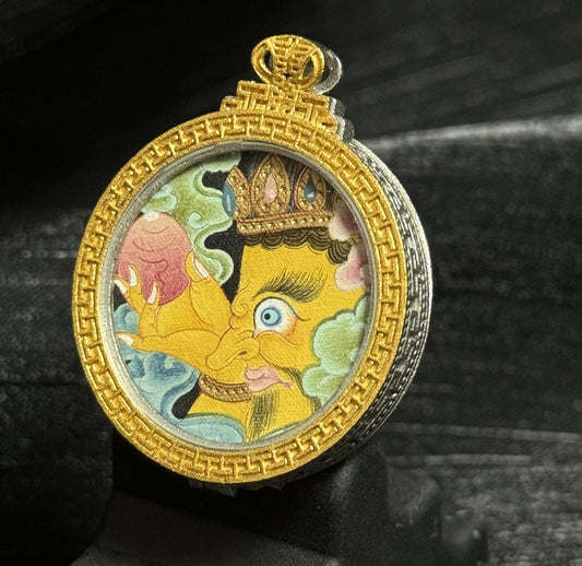 Angled view of the Yellow Jambhala mini thangka pendant showing detailed mineral pigment painting, golden pendant frame, and textured silver protective case surrounding the circular artwork.