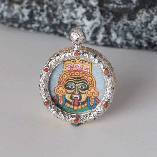 Front view of a Zaki Lhamo mini thangka pendant, hand-painted with coarse mineral pigments, set in a round jewelry-grade copper case with ornamental detailing.