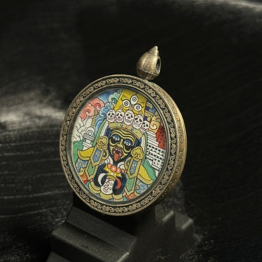Angled view of Zaki Lhamo round mini thangka pendant showing detailed mineral pigment painting, expressive deity portrait, and engraved circular metal frame.