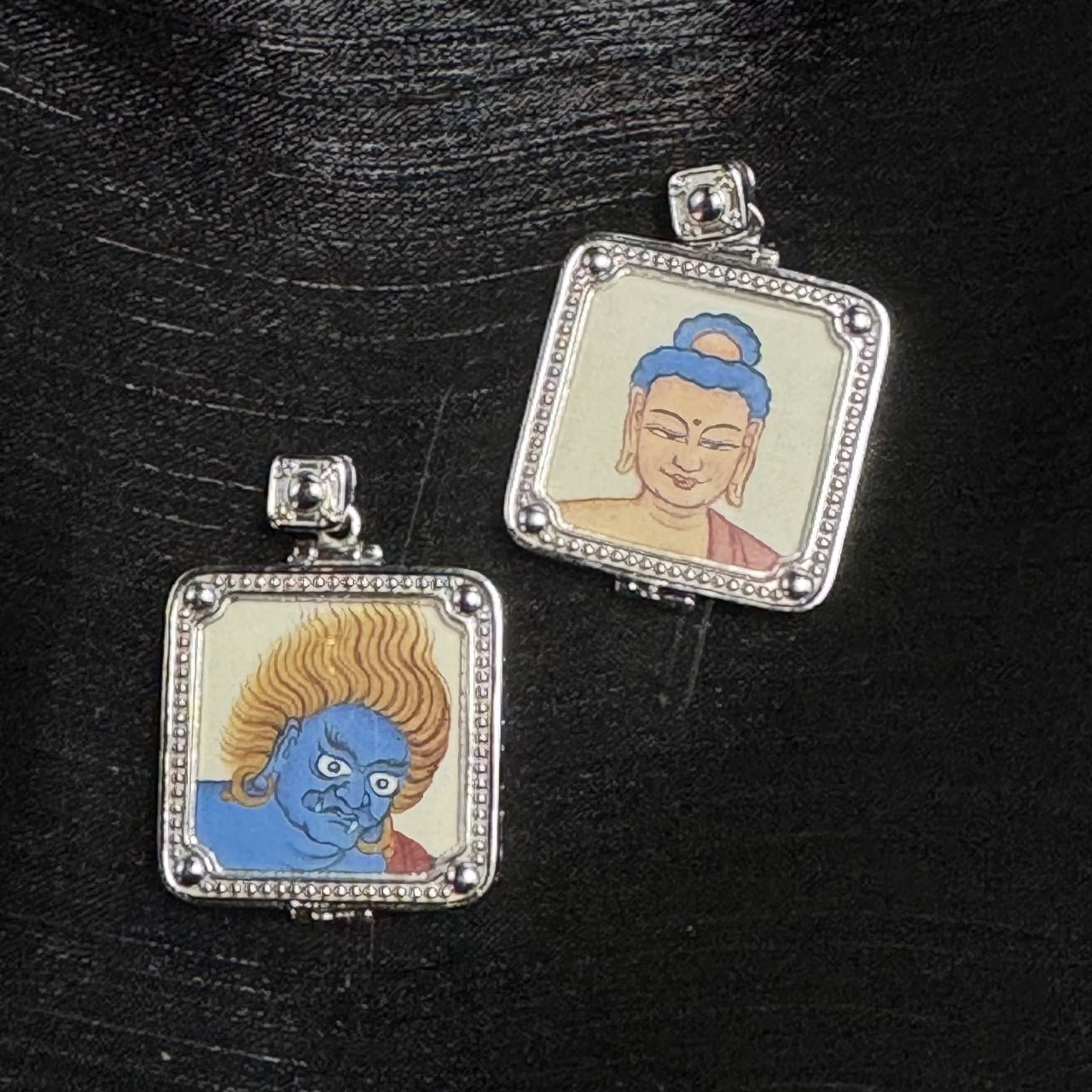 Two square mini thangka pendants on black background: Acalanatha portrait with blue face and flame-like yellow hair, and Amitabha Buddha portrait with calm face and blue hair bun, both in silver-tone frames with dotted edging, round corner beads, and a square top bail