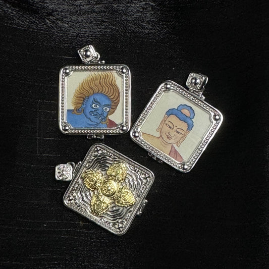 Three square pendants on a black textured surface: two show single-sided hand-painted portraits of Acalanatha (blue face with flame-like hair) and Amitabha Buddha (calm face with blue hair bun), each in a silver-tone square frame with a dotted border and round corner beads, plus one pendant showing a raised gold-tone vajra motif on the back