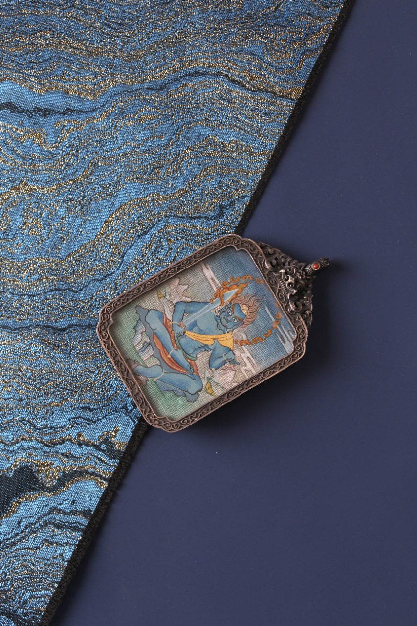 Angled view of an Acalanatha (Fudo Myo-o) mini thangka pendant in an ornate vintage silver gawu frame, showing the hand-painted thangka artwork against a deep blue backdrop.