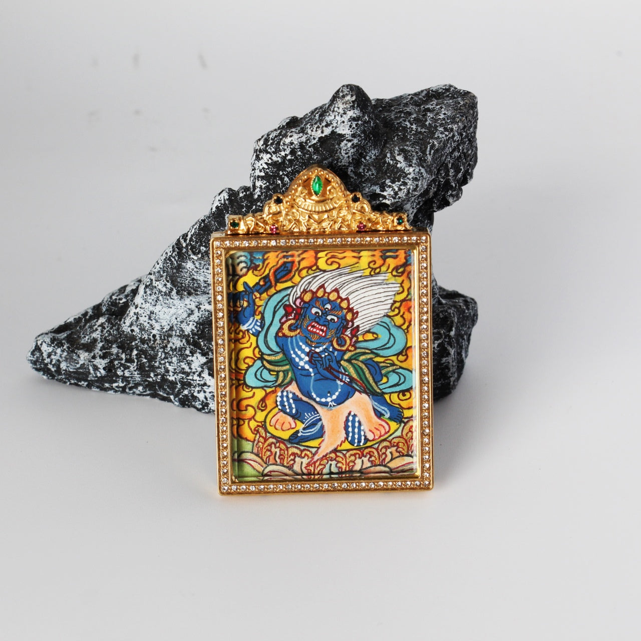 Angled view of Acalanatha (Fudo Myo-o) mini thangka pendant showing the hand-painted blue wrathful deity figure with flame background, gold-tone rectangular frame with crystal border, and ornate crown top with a green gemstone, photographed on a rock prop.