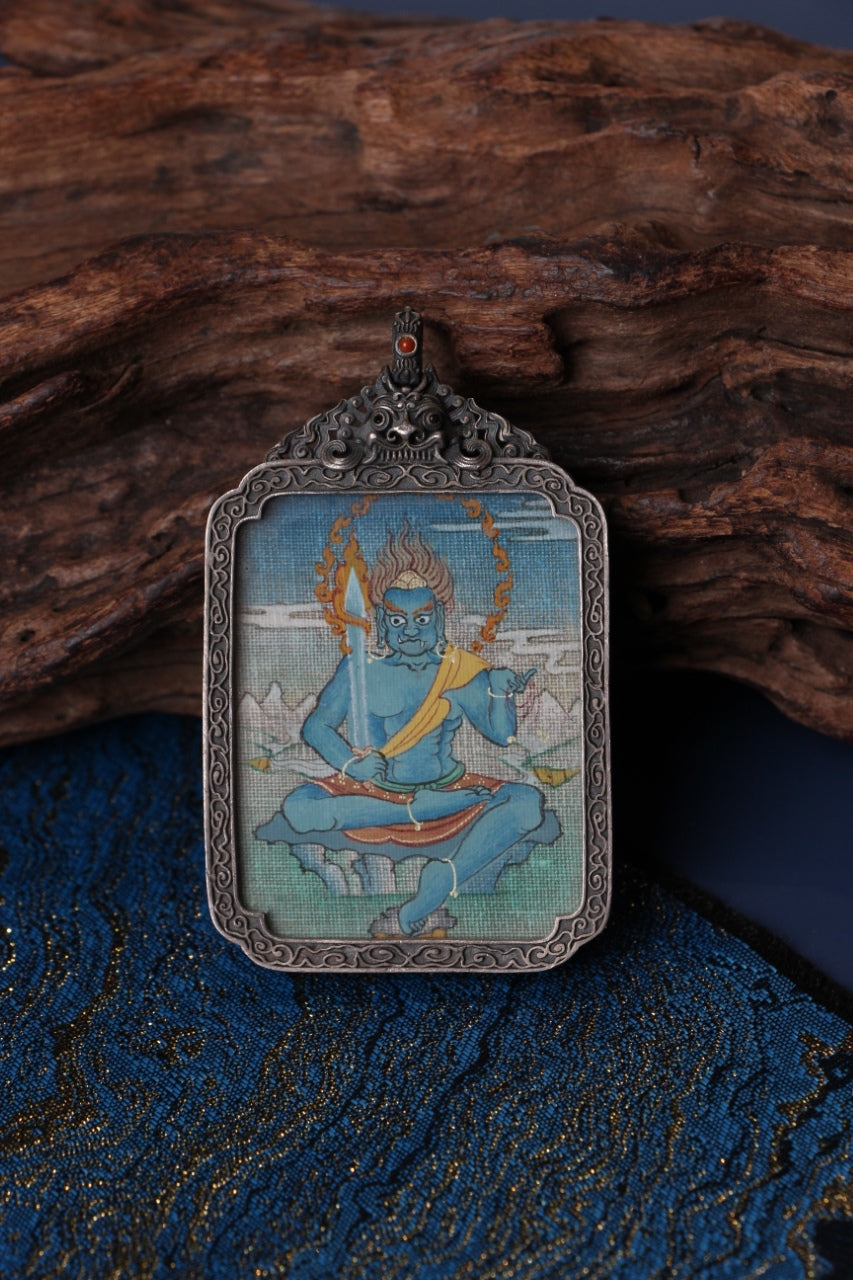 Close-up of an Acalanatha (Fudo Myo-o) mini thangka pendant, hand-painted with mineral pigments and framed in a detailed vintage silver gawu box, photographed on a wooden surface with blue fabric.
