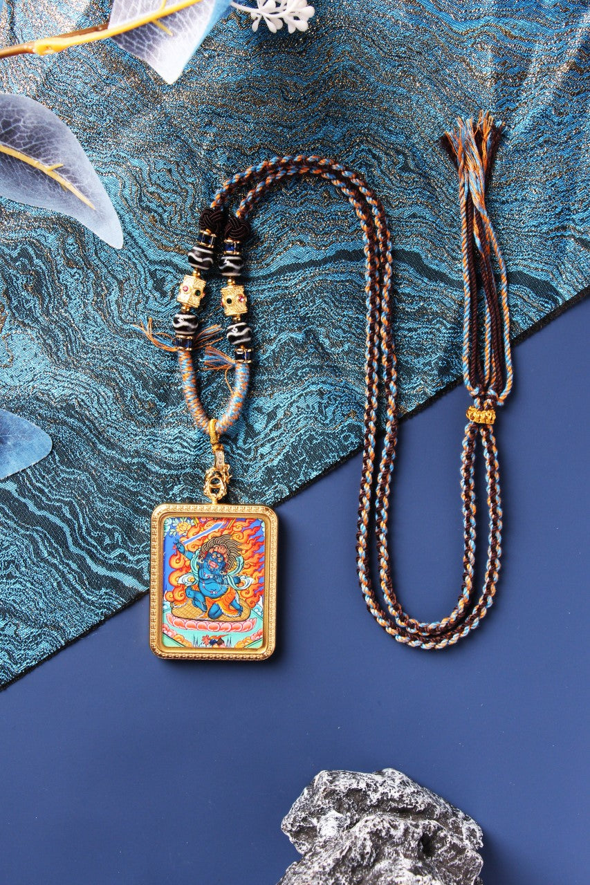 Full layout photo showing the Acalanatha mini thangka pendant in a gold-tone titanium steel case with a long braided cord, adjustable tassel end, and decorative bead segments.