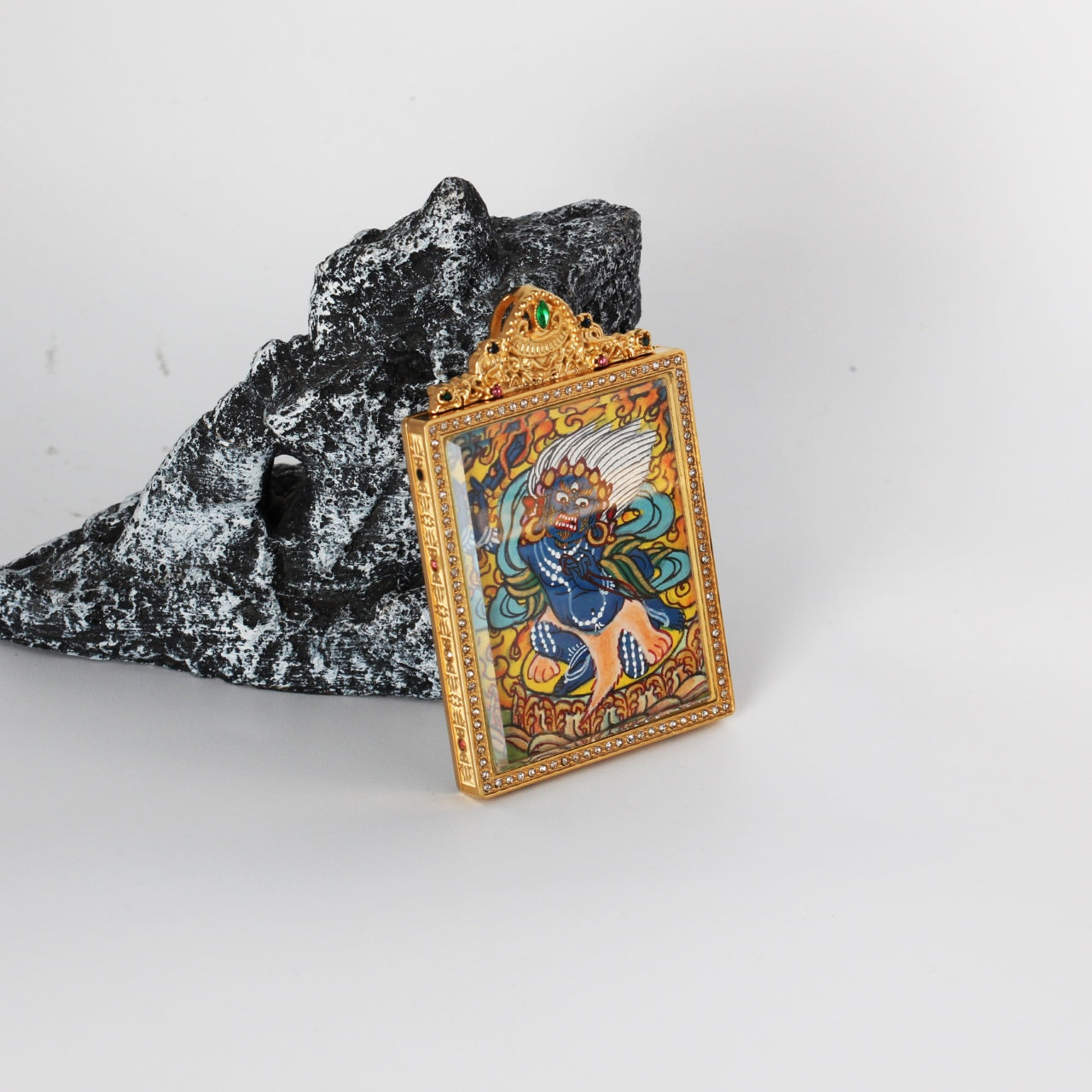 Side-angled photo of Acalanatha (Fudo Myo-o) mini thangka pendant showing the gold-tone titanium steel case profile, crystal border around the front window, and the hand-painted wrathful deity artwork inside.