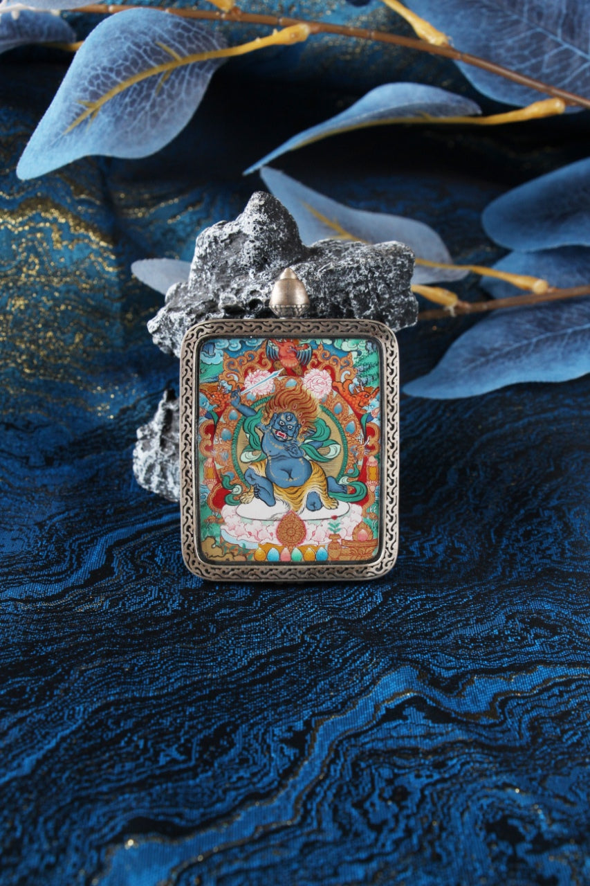 Front view of an Acalanatha (Fudo Myo-o) Menri-style mini thangka pendant, hand-painted in mineral pigments and set in a silver gawu case on blue fabric with stone.