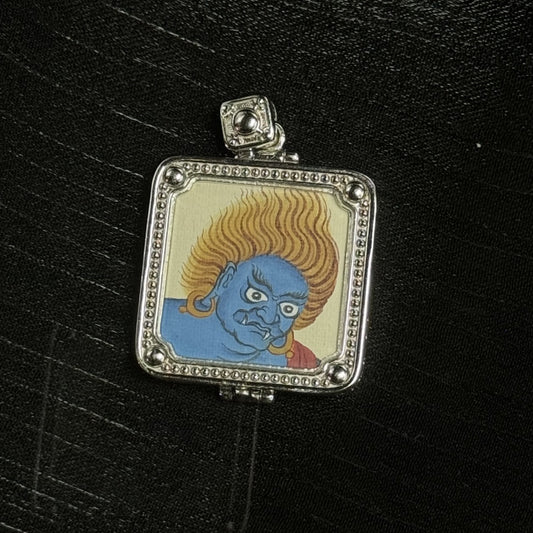Close-up of Acalanatha mini thangka pendant showing a blue-faced portrait with intense expression and flame-like yellow hair, inside a silver-tone square frame with dotted border, round corner beads, and a square top bail
