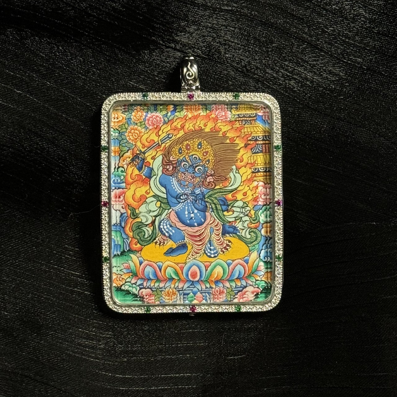 Detailed close-up of the Acalanatha mini thangka pendant highlighting the vibrant mineral pigment painting, fierce expression of Fudo Myo-o, flame background, and ornate lotus base inside a silver pendant setting.