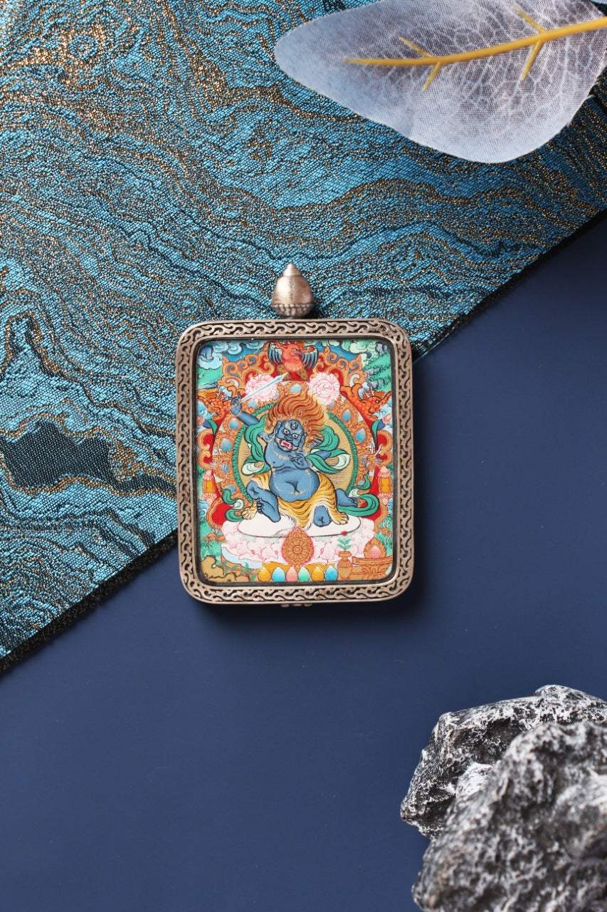 Front display of an Acalanatha (Fudo Myo-o) Menri-style mini thangka pendant in a silver gawu case, photographed with blue-gold brocade and rock props.